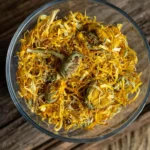 A glass bowl filled with vibrant dried calendula flowers, showcasing their bright orange and yellow petals mixed with green stems, placed on a rustic wooden surface.