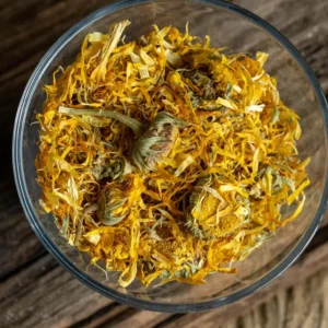 A glass bowl filled with vibrant dried calendula flowers, showcasing their bright orange and yellow petals mixed with green stems, placed on a rustic wooden surface.