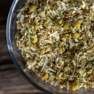 A close-up view of a glass bowl filled with dried chamomile flowers, featuring delicate white petals and yellow centers, scattered with small stems and leaves, placed on a dark wooden surface.