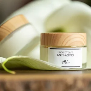 A small jar of face cream labeled "Face Cream Anti-Aging AV" with a wooden lid sits on a large white petal with a green tip, placed on a wooden surface, with another jar partially visible in the blurred background.