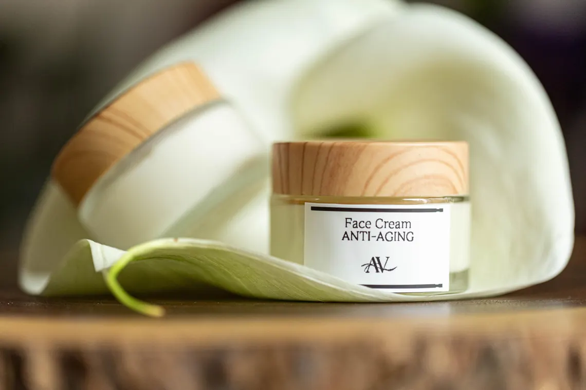 Organic Face Cream Age-Fighting |Handcrafted Skincare | Natural Gift for Her 1 A small jar of face cream labeled "Face Cream Anti-Aging AV" with a wooden lid sits on a large white petal with a green tip, placed on a wooden surface, with another jar partially visible in the blurred background.