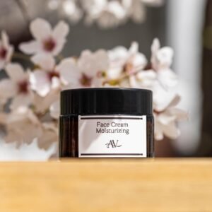 A small dark jar of face cream labeled "Face Cream Moisturizing AV" sits on a wooden surface, with a blurred background of white flowers with pink centers, creating a soft and serene setting.