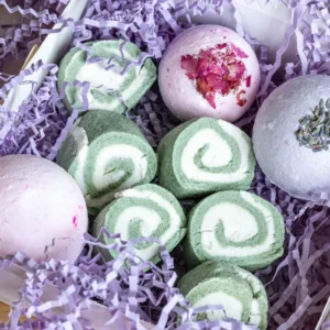 A collection of handmade bubble bars and bath bombs arranged on a white marble surface. The bubble bars are pastel pink and blue with a creamy texture, while the bath bombs are round, white with speckles of dried flowers, and one has a light purple hue with lavender buds. A few dried flower petals are scattered around, with a soft, blurred background of a bathroom setting.