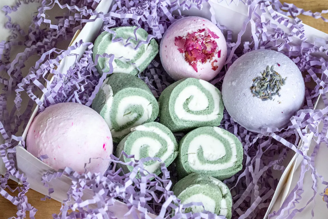 Handmade Bubble Bars and Bath Bombs for Relax Organic Skincare 1 A collection of handmade bubble bars and bath bombs arranged on a white marble surface. The bubble bars are pastel pink and blue with a creamy texture, while the bath bombs are round, white with speckles of dried flowers, and one has a light purple hue with lavender buds. A few dried flower petals are scattered around, with a soft, blurred background of a bathroom setting.
