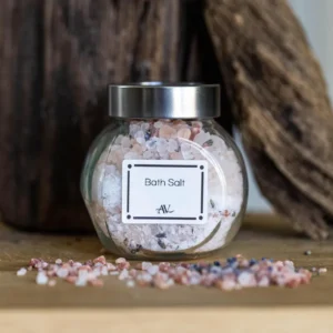 A glass jar filled with lavender bath salts mixed with essential oils, labeled "Bath Salt AdoreVera," sits on a wooden surface with a rustic wooden background. Some of the pink and white bath salts with lavender buds are scattered around the jar, emphasizing its natural, relaxing vibe.