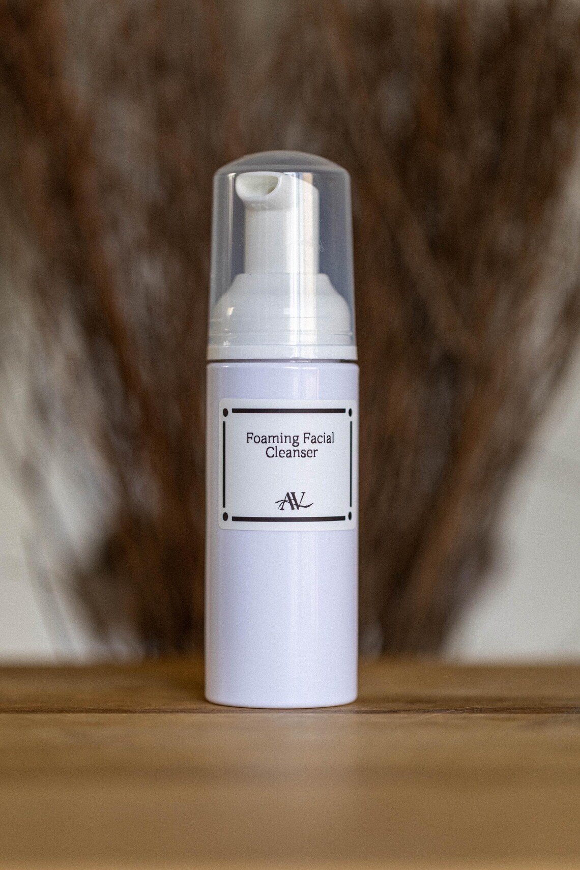 Orange Cleansing Face Wash 1 A white plastic bottle of orange cleansing face wash with a foamer pump and a clear cap, labeled "Foaming Facial Cleanser" with the initials "AV," is placed on a wooden surface with a blurred brown background.