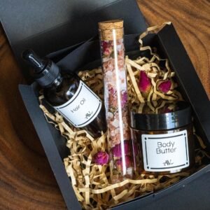 A black gift box with shredded straw filler contains three skincare products: a bottle of hair oil with a dropper, a glass tube of bath salts with dried rose petals, and a jar of body butter. The products are labeled with white stickers and black text, surrounded by dried pink rose petals on a wooden surface.