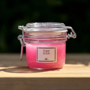 A glass jar of Organic Raspberry Sugar Scrub, labeled "Raspberry Scrub AV," with a metallic lid, sits on a white linen cloth on a wooden surface. Scattered around the jar are dried raspberries and pink sugar grains, with a blurred background of soft pink flowers and greenery.