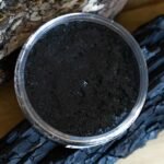 A glass jar filled with a dark, grainy organic salt scrub made with Black Hawaiian Salt and Dead Sea Salt, placed on a wooden surface alongside pieces of charcoal and a rough log, creating a rustic, natural setting.