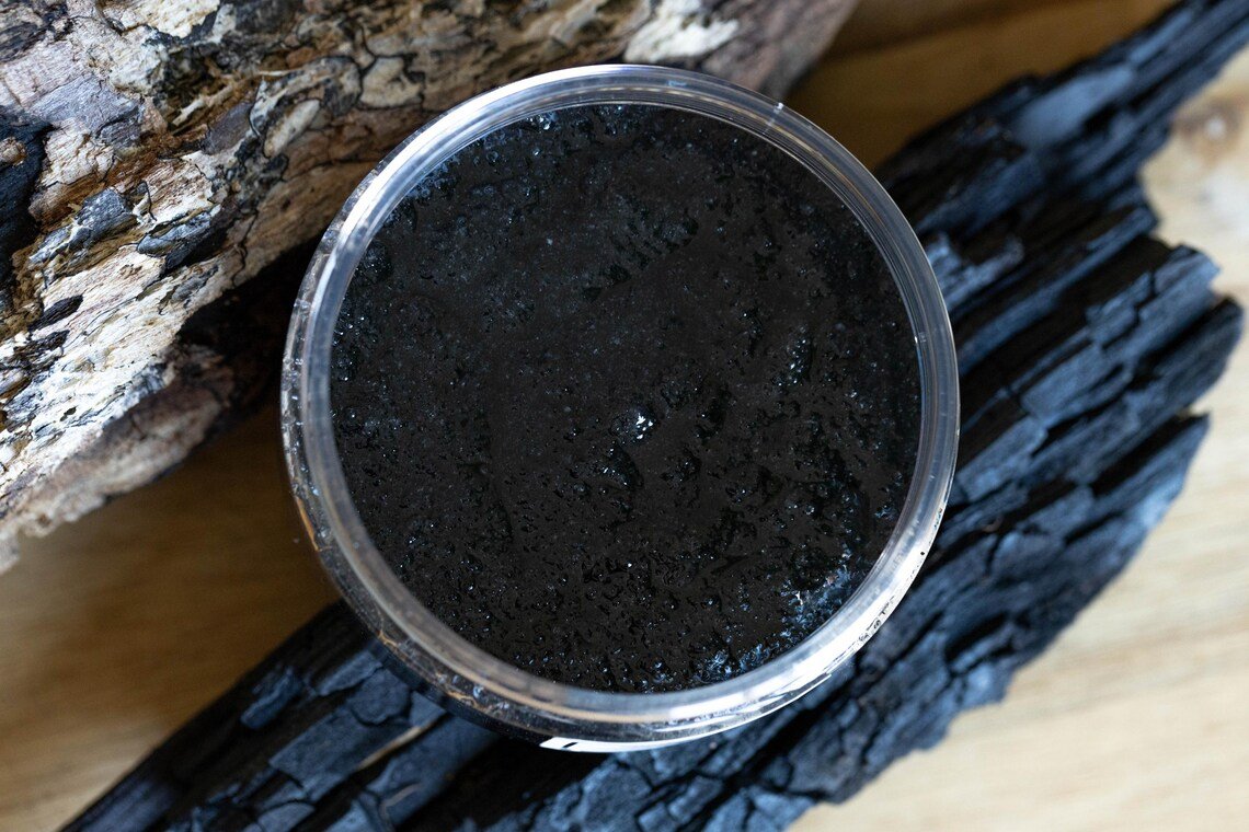 Organic Salt Scrub With Black Hawaiian Salt and Dead Sea Salt 1 A glass jar filled with a dark, grainy organic salt scrub made with Black Hawaiian Salt and Dead Sea Salt, placed on a wooden surface alongside pieces of charcoal and a rough log, creating a rustic, natural setting.