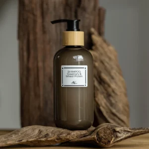 A frosted brown bottle of organic shampoo labeled "Shampoo Rosemary & Wheat Protein AV" with a wooden cap and black pump, placed on a piece of weathered wood against a rustic wooden background, highlighting its natural aesthetic.