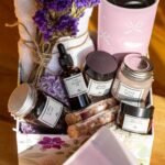 A gift box filled with self-care items, including a pink tumbler, a white towel with dried purple flowers tied with twine, and various skincare products labeled "Hair Growth Oil," "Body Butter," "Facial Mask," "Coffee Scrub," and "Face Cream." The box is decorated with purple shredded paper and sits on a wooden surface with sunlight streaming in.