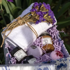 An Organic Spa Gift Box for Her, featuring a wooden box filled with skincare products like a frosted glass jar of face cream, a bottle of body oil, and a lavender bath bomb, all nestled in beige shredded paper. The box is placed on a white linen cloth on a wooden surface, with dried lavender sprigs and a rolled white towel in the background, creating a serene spa vibe.