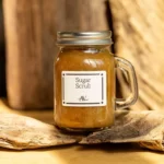 A glass jar with a handle, filled with a golden-brown organic sugar body scrub, labeled "Sugar Scrub AV," placed on a piece of weathered wood against a rustic wooden background.
