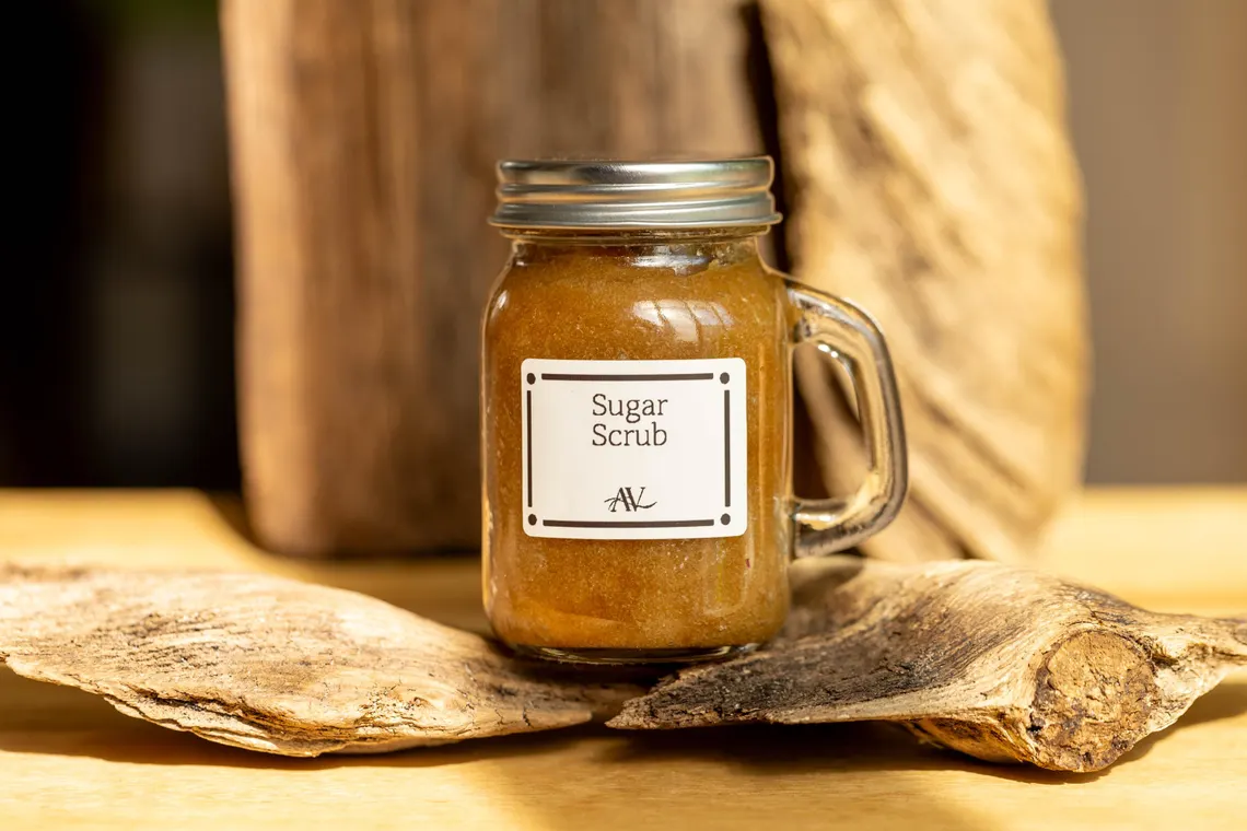 Organic Sugar Body Scrub | Exfoliating Spa Gift for Her Selfcare 1 A glass jar with a handle, filled with a golden-brown organic sugar body scrub, labeled "Sugar Scrub AV," placed on a piece of weathered wood against a rustic wooden background.