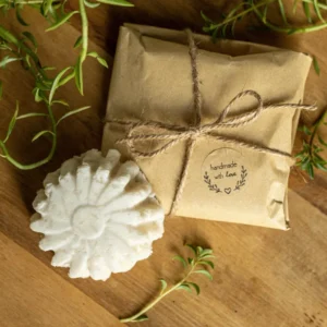 A white, flower-shaped solid handmade shampoo bar sits beside a kraft paper package tied with twine on a wooden surface. The package has a circular label that reads "handmade with love," surrounded by green leafy sprigs.