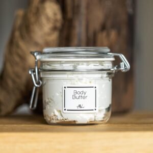 A small glass jar of Whipped Shea Body Butter, labeled "Shea Body Butter AV," with a metallic lid, sits on a smooth wooden surface. The jar is surrounded by small chunks of raw shea butter and a soft beige linen cloth, with a blurred background of warm, neutral tones.