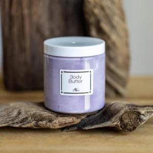 A small glass jar of lavender body butter, labeled "Body Butter AdoreVera," with a wooden lid, sits on a smooth stone surface. Surrounding the jar are dried lavender sprigs and a soft white linen cloth, with a blurred background of a purple-toned spa setting.