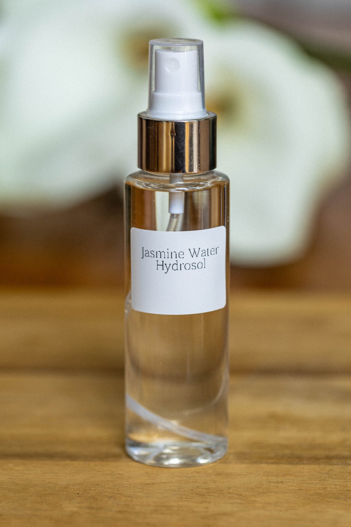 A clear glass spray bottle labeled "Jasmine Water Hydrosol" sits on a wooden surface. The bottle has a metallic cap and white spray nozzle, with a soft-focus background of blurred white flowers, creating a serene and natural atmosphere.