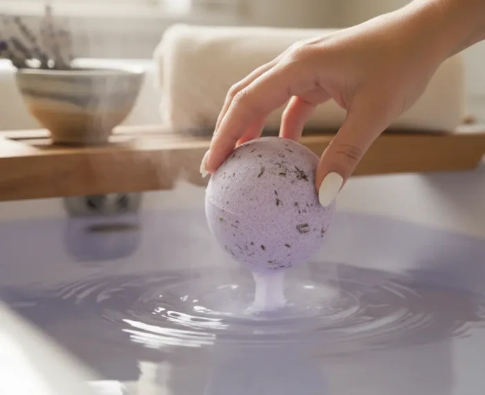 Hand gently placing a natural lavender bath bomb into steaming water with soft light and botanicals.